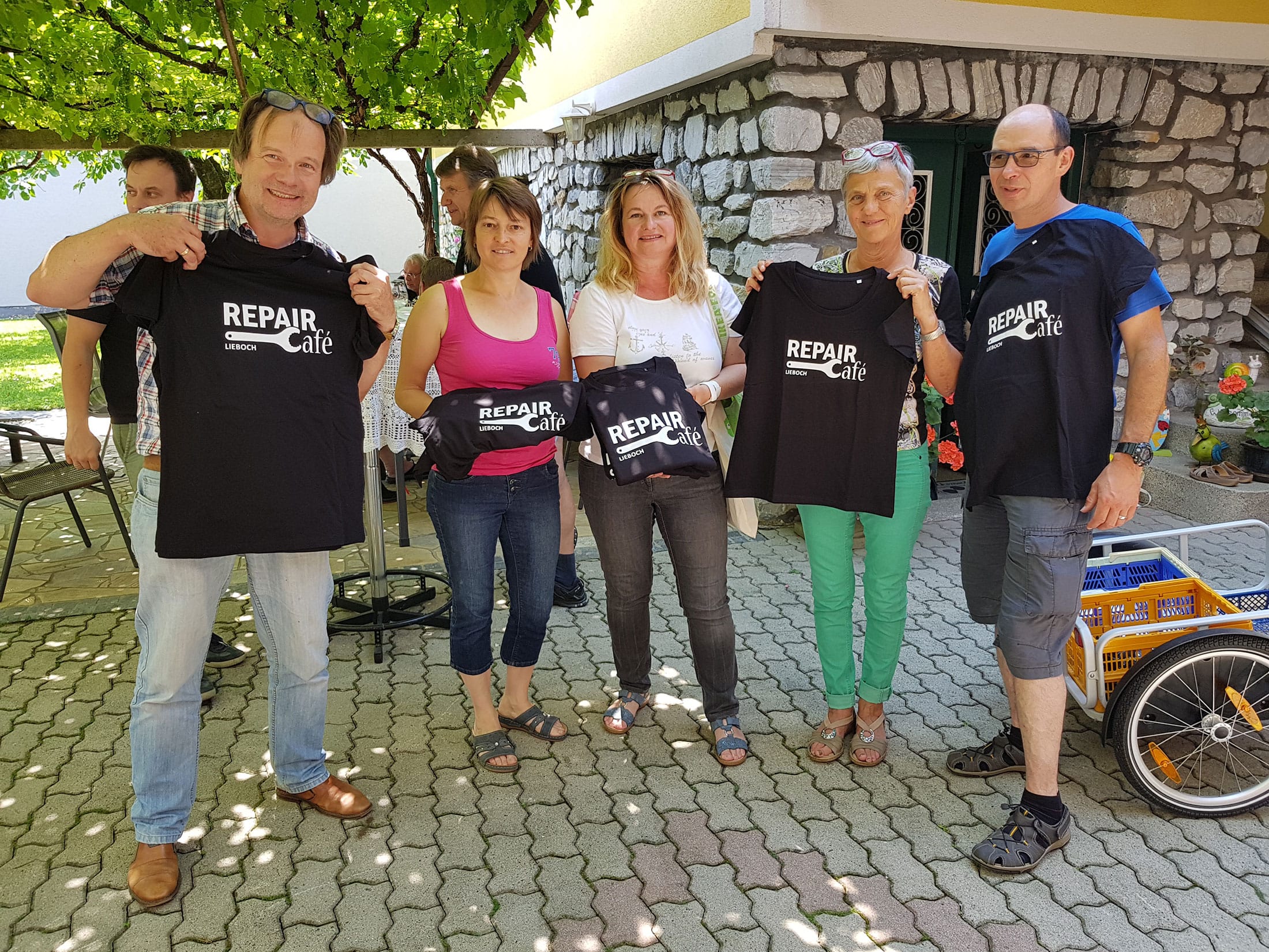 5 Personen stehen vor einem steinernen Gebäude und halten schwarze T-Shirts mit dem Aufdruck 'Repair Café' in die Kamera