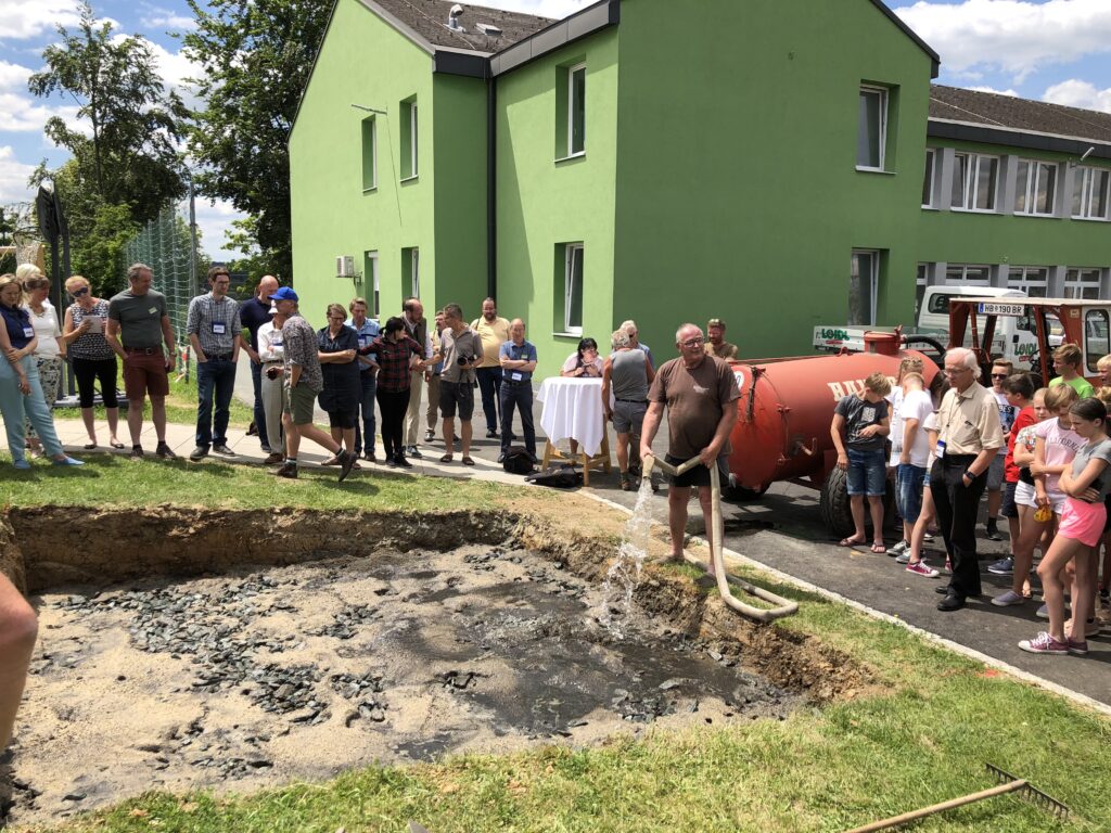 Ein Klimabaum wird bei der VS Kaindorf gesetzt. Ein Mann füllt hierfür ein ausgehobenes Becken in der Wiese und die Zuschauer sehen zu.