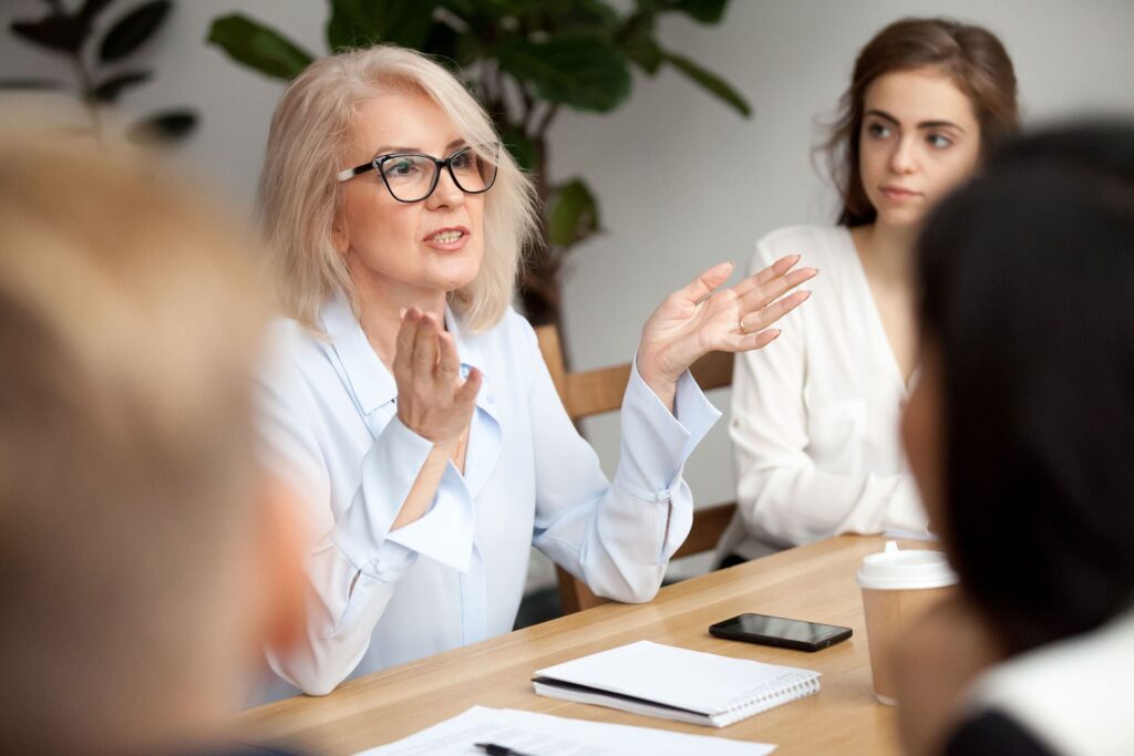 Eine Dame in weißer Bluse diskutiert mit drei weiteren Personen in einer Runde auf einem Arbeitstisch, vor ihr liegen Unterlagen