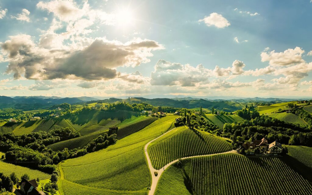 Blick über begrünte Berge, die Sonne scheint am leicht bewölkten, blauen Himmel